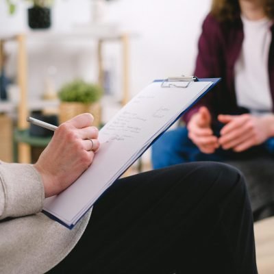 Close-up of a therapist writing notes on a clipboard while conversing with a patient.